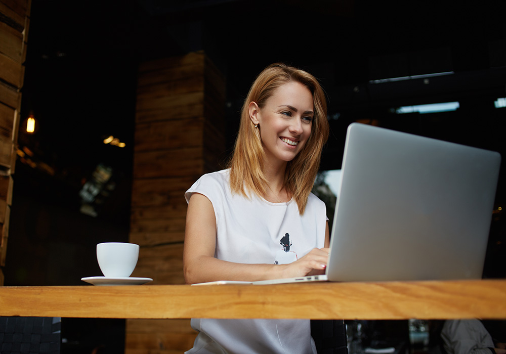 A woman smiling at her laptop in a cafe, with a cup of coffee next to her.